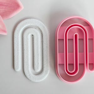 Op de afbeelding: Twee abstracte, boogvormige objecten. Eén is wit met een textuuroppervlak, de andere is roze en transparant. Een roze bloemblaadje bevindt zich in de linkerbovenhoek. De objecten staan op een wit oppervlak.