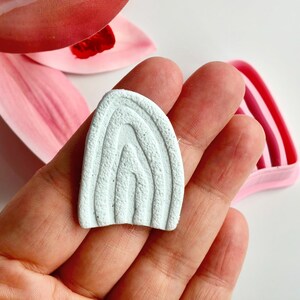 Op de afbeelding: Een lichtblauw, getextureerd, boogvormig kleistuk met gelaagde groeven, vastgehouden in een hand. Roze bloemblaadjes en een roze en rood gestreept object bevinden zich op de achtergrond.