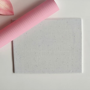 Può includere: Un mattarello cilindrico rosa chiaro con una consistenza a coste posizionato diagonalmente su un foglio di cartoncino bianco rettangolare caratterizzato da un fine motivo a righe orizzontali e piccole macchie scure.