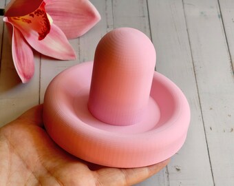 May include: A light pink, round object with a cylindrical top, held in a hand. The object appears to be a decorative item or a small sculpture. The background includes a pink orchid flower and a light-colored wooden surface.