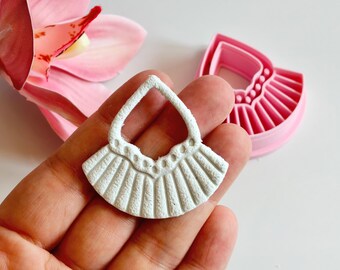 May include: A white, textured earring pendant with a fan-like design, held in a hand. A pink earring cutter is next to the pendant. A pink orchid flower is in the background.