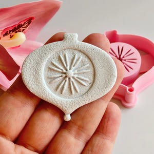 Könnte beinhalten: Weißer, strukturierter Ausstecher in Ornamentform mit Sternmuster. Ein rosa Ausstecher und eine rosa Orchideenblüte befinden sich im Hintergrund. Das Ornament hat oben eine Schlaufe und unten eine kleine Tropfenform.