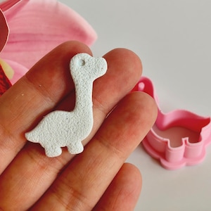 Op de afbeelding: Een klein, lichtgekleurd, dinosaurusvormig knutselstuk wordt in een hand gehouden, naast een roze, dinosaurusvormige koekjesuitsteker van plastic. Het knutselstuk heeft een textuuroppervlak en een gat bij de kop.