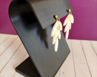 May include: A pair of white and gold leaf-shaped earrings displayed on a black earring stand. The earrings have a gold-colored post and a marbled design. The stand is angled and has a curved top.