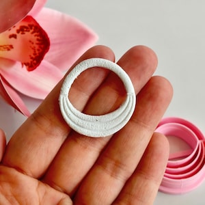 Può includere: Un utensile artigianale circolare bianco e strutturato con un design curvo decorativo. Sono visibili anche un fiore rosa e una serie di utensili circolari rosa. Gli strumenti sembrano essere per l'artigianato o la cottura.