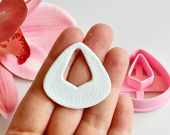 May include: A white, textured teardrop-shaped earring component is held in a hand, next to a pink plastic earring cutter. A pink orchid flower is in the background. The earring component has a textured surface.
