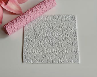 May include: A pink textured rolling pin and a white square with a floral pattern. The rolling pin is a light pink color and has a repeating floral design. The white square has a raised floral pattern.