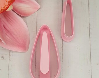 May include: Two pink teardrop-shaped cookie cutters, one large and one small, sit on a white wooden surface. The cutters have a raised inner shape. A pink flower petal is visible in the upper left corner.