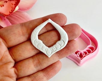 May include: A white, teardrop-shaped cookie cutter with a decorative floral design along the bottom edge. A pink cookie cutter of the same shape is also visible. The items are held in a hand.