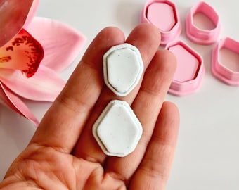 May include: Two white, geometric-shaped clay pieces with a textured border are held in a hand. Several pink, geometric-shaped cookie cutters are in the background. A pink orchid flower is in the upper left corner.
