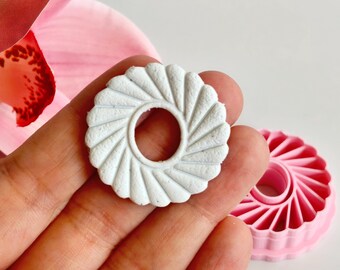 May include: A white, circular cookie cutter with a fluted edge and a central hole is held in a hand. A pink cookie cutter of the same design is in the background. A pink orchid flower is in the upper left corner.