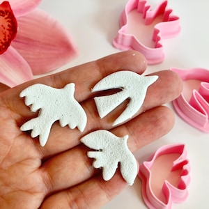 Op de afbeelding: Drie witte duifvormige kleisnijders met een textuuroppervlak worden in een hand gehouden, naast roze plastic duifvormige koekjessnijders. Op de achtergrond is een roze bloem te zien.