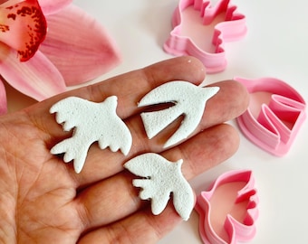 May include: Three white dove-shaped clay cutouts with a textured surface are held in a hand, alongside pink plastic dove-shaped cookie cutters. A pink flower is visible in the background.