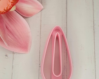 May include: A pink plastic teardrop-shaped cookie cutter with a central vertical divider. The cutter is set against a light wood-grain background, with a pink flower petal in the upper left corner.