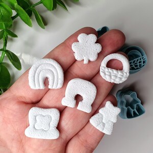 Może przedstawiać: Zbiór białych, teksturowanych kształtów z gliny w różnych wzorach, w tym tęcza, koniczyna, kosz i gwiazda. Kształty są trzymane w dłoni, z niebieskimi plastikowymi wykrawaczkami w tle. Do projektów rzemieślniczych lub piekarniczych.