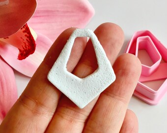 May include: A light blue, textured, geometric earring component is held in a hand, with a pink flower and a pink earring cutter in the background. The earring component has a unique, open design.