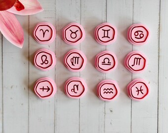 May include: A collection of twelve pink, hexagonal-shaped objects, each bearing a different zodiac symbol in dark red. The objects are arranged on a light wood-grain surface, with a pink flower in the upper left corner.