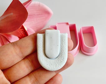 May include: A light blue, U-shaped clay piece with a white rectangular top, held in a hand. Two pink plastic cutters are in the background, along with a pink flower.