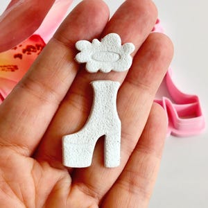 Puede incluir: Primer plano de una mano que sostiene dos objetos blancos pequeños y texturizados: una forma de nube y una bota de plataforma. Una flor rosa y un objeto en forma de zapato rosa están en el fondo. Los objetos parecen ser de arcilla.