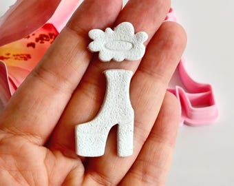 May include: A close-up of a hand holding two small, textured, white objects: a cloud shape and a platform boot. A pink flower and a pink shoe-shaped object are in the background. The objects appear to be made of clay or a similar material.