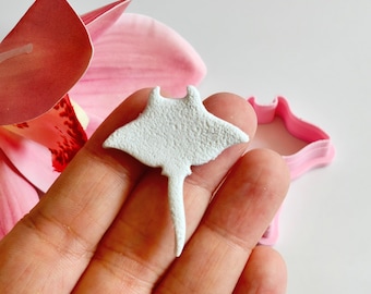 May include: A small, textured, white stingray-shaped craft element is held in a hand. A pink flower and a pink stingray-shaped cookie cutter are in the background. The craft element has a textured surface and a long tail.