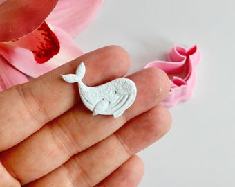 May include: A small, light blue whale-shaped craft element with star details is held in a hand. A pink whale-shaped cookie cutter is in the background. The craft element has a textured surface and is likely made of clay or fondant.
