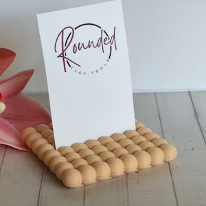 Op de afbeelding: Een rechthoekige beige standaard met een raster van kleine, verhoogde bolletjes op het oppervlak, ontworpen om een witte kaart vast te houden. De kaart bevat een cirkelvormig logo met de tekst Rounded Clay Tools in donkerpaarse en zwarte letters.