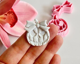May include: A white, pumpkin-shaped craft with a bow, held in a hand. Pink pumpkin and bow-shaped cookie cutters are in the background, along with a pink orchid flower. The pumpkin has carved details.
