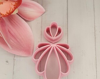 May include: Two pink, flower-shaped cookie cutters. One is a larger, multi-petal design, and the other is a smaller, simpler version. The cutters are made of plastic and are set against a white wooden background, with a pink flower in the background.