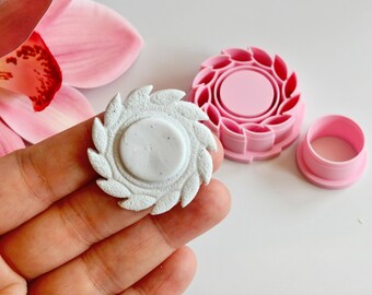 May include: A white, circular clay piece with a textured, leaf-like border is held in a hand. Beside it are two pink plastic cookie cutters, one with a similar leaf design and a circular center, and a smaller, round cutter. A pink orchid flower is in the background.