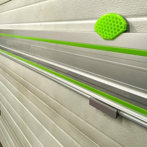 Op de afbeelding: Een zilveren metalen rail met groene tape en een grijze plastic stop. De rail is gemonteerd op een witte garagedeur.