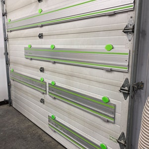 Op de afbeelding: Vier zilveren en groene railsagen zijn gemonteerd op een witte garagedeur. Elke zaag heeft een groene handgreep en een groene en zilveren rail. De zagen zijn gelabeld met de tekst "Festool" en "TS 55 REQ" in zwart.