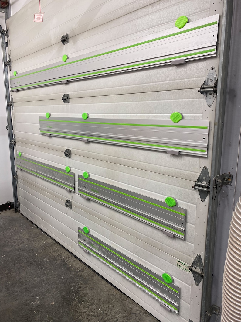 Op de afbeelding: Vier zilveren en groene railsagen zijn gemonteerd op een witte garagedeur. Elke zaag heeft een groene handgreep en een groene en zilveren rail. De zagen zijn gelabeld met de tekst "Festool" en "TS 55 REQ" in zwart.