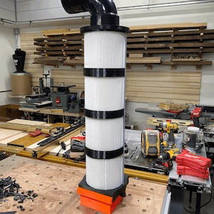 May include: A white filter with black bands, attached to a black plastic pipe, sits on a wooden workbench. The filter is connected to a black plastic pipe with a 90-degree bend. The filter is surrounded by tools and other equipment in a workshop.