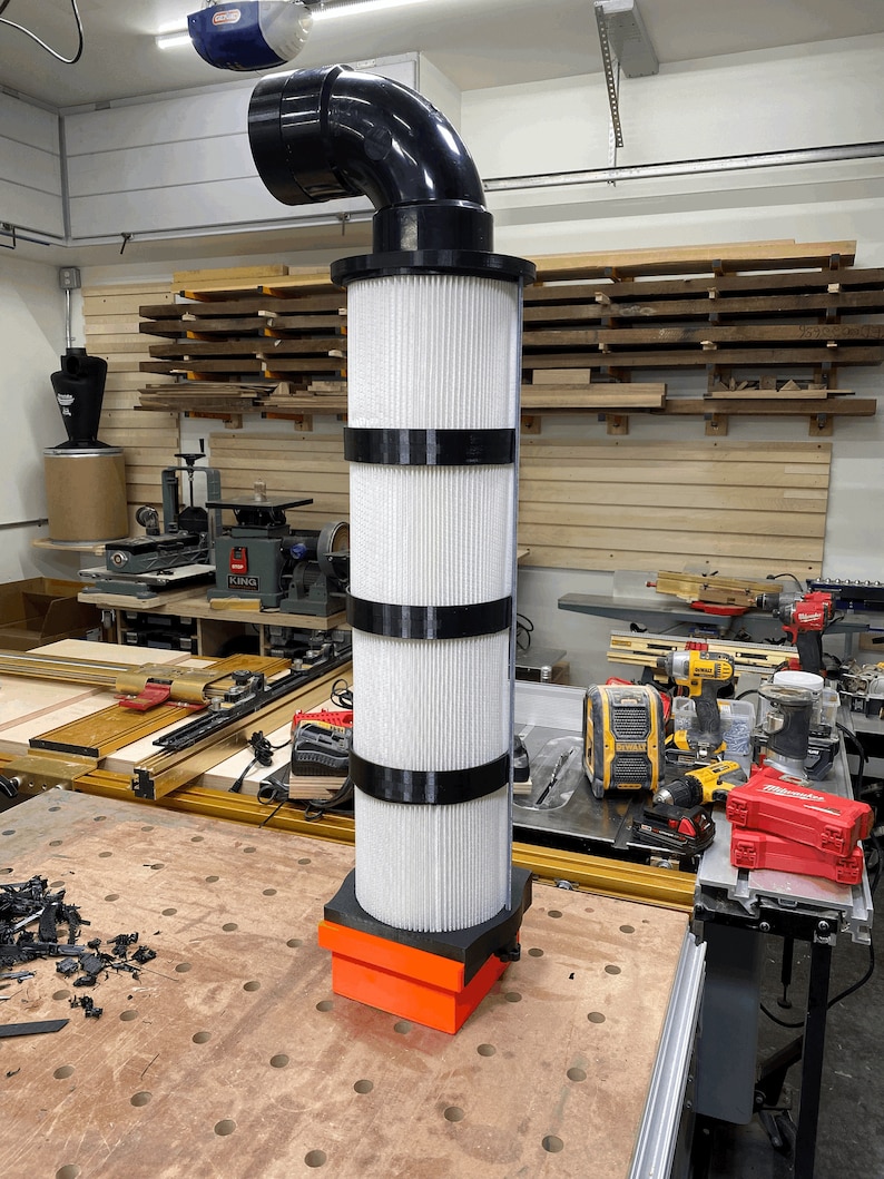 May include: A white filter with black bands, attached to a black plastic pipe, sits on a wooden workbench. The filter is connected to a black plastic pipe with a 90-degree bend. The filter is surrounded by tools and other equipment in a workshop.