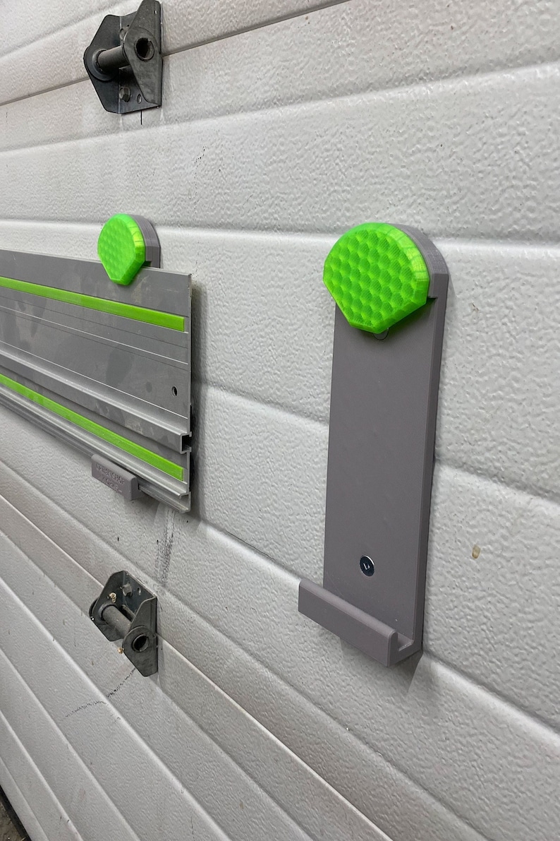 Op de afbeelding: Een grijze wandbevestiging met een groene, honingraatvormige knop. De wandbevestiging heeft een klein plankje aan de onderkant. Er is een tweede groene knop op de muur links van de wandbevestiging.