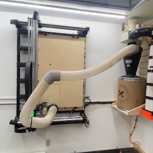 May include: A white dust collection system with a flexible hose connected to a large machine. The system is mounted on a wall and has a large, cylindrical container with a brown paper bag. The bag has a logo that says "The Wood Whisperer".