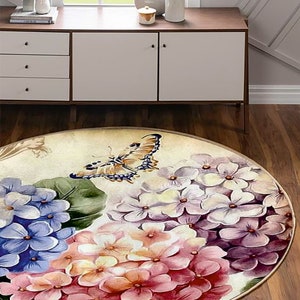 Puede incluir: Alfombra redonda con un diseño floral que presenta una mariposa y hortensias en tonos de rosa, morado y azul. La alfombra tiene un fondo beige y un borde dorado.