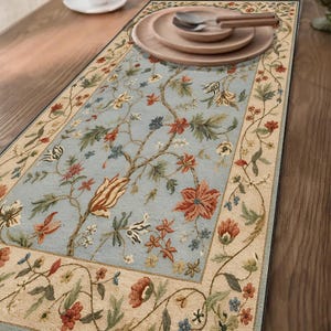 Puede incluir: Un camino de mesa rectangular con un panel central azul claro que presenta un patrón floral extendido en tonos rojo, naranja, verde y blanco. El camino tiene un borde ancho de color crema decorado con un motivo continuo de enredaderas y flores.
