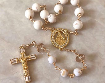 May include: A gold-toned rosary with white marble-like beads. The rosary features a gold crucifix and a circular pendant with a religious figure. The beads are connected by gold wire.
