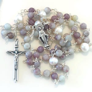 May include: A rosary with a silver crucifix and a silver Virgin Mary pendant. The beads are a mix of lavender, gray, and white, with a few large white pearl beads. The beads are strung on silver wire.