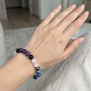 Könnte beinhalten: Ein Perlenarmband mit einer Abfolge von runden, polierten Steinen in den Farben Dunkelviolett, glänzendem Schwarz, blassem Rosa, Metallic-Grau und gesprenkeltem Blau, die auf einem flexiblen elastischen Band aufgezogen sind.