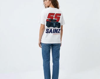 May include: A white t-shirt with a graphic on the back featuring the number "55" in red, a race car, and the name "SAINZ" in blue. The shirt is paired with blue jeans and white sneakers.
