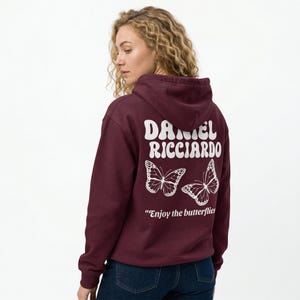 May include: A maroon hoodie with white text and butterfly graphics. The back of the hoodie features the name "DANIEL RICCIARDO" above two butterfly illustrations and the phrase "Enjoy the butterflies."