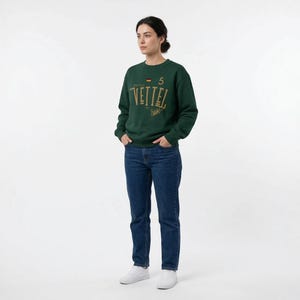 May include: A dark green sweatshirt with the name "VETTEL" in gold lettering. The number "5" and a small German flag are above the name. The person is wearing blue jeans and white sneakers.