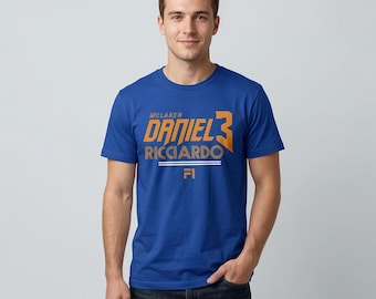 May include: A blue t-shirt featuring the name "DANIEL 3 RICCIARDO" in orange, with "McLaren" above and "FI" below. The shirt is short-sleeved and worn by a person.