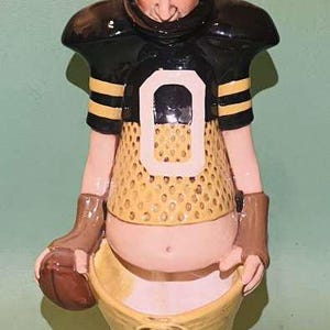 May include: A ceramic figurine of a football player wearing a black and gold uniform with a large 'O' on the front. The figurine is holding a football and has a comical expression on its face.