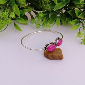 Könnte beinhalten: Ein silbernes Armreif-Armband mit zwei rosa ovalen Edelsteinen. Das Armband liegt auf einem braunen Stein.