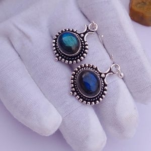 May include: A pair of silver earrings featuring oval-shaped labradorite gemstones with blue iridescence. Each stone is set in a decorative silver frame with a beaded edge and a small loop at the top for attachment, showcasing a metallic finish.