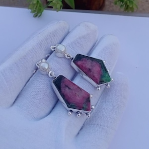May include: A pair of dangle earrings featuring polished, irregular-shaped ruby zoisite stones set in silver metal. Each earring is topped with a small, round white pearl and accented with tiny silver spheres along the bottom edge of the stone setting.